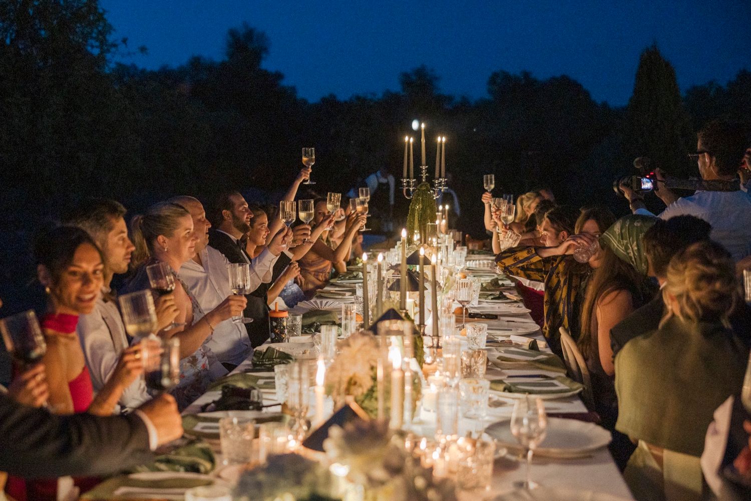 Candlelit wedding dinner table with guests under dark blue evening sky.