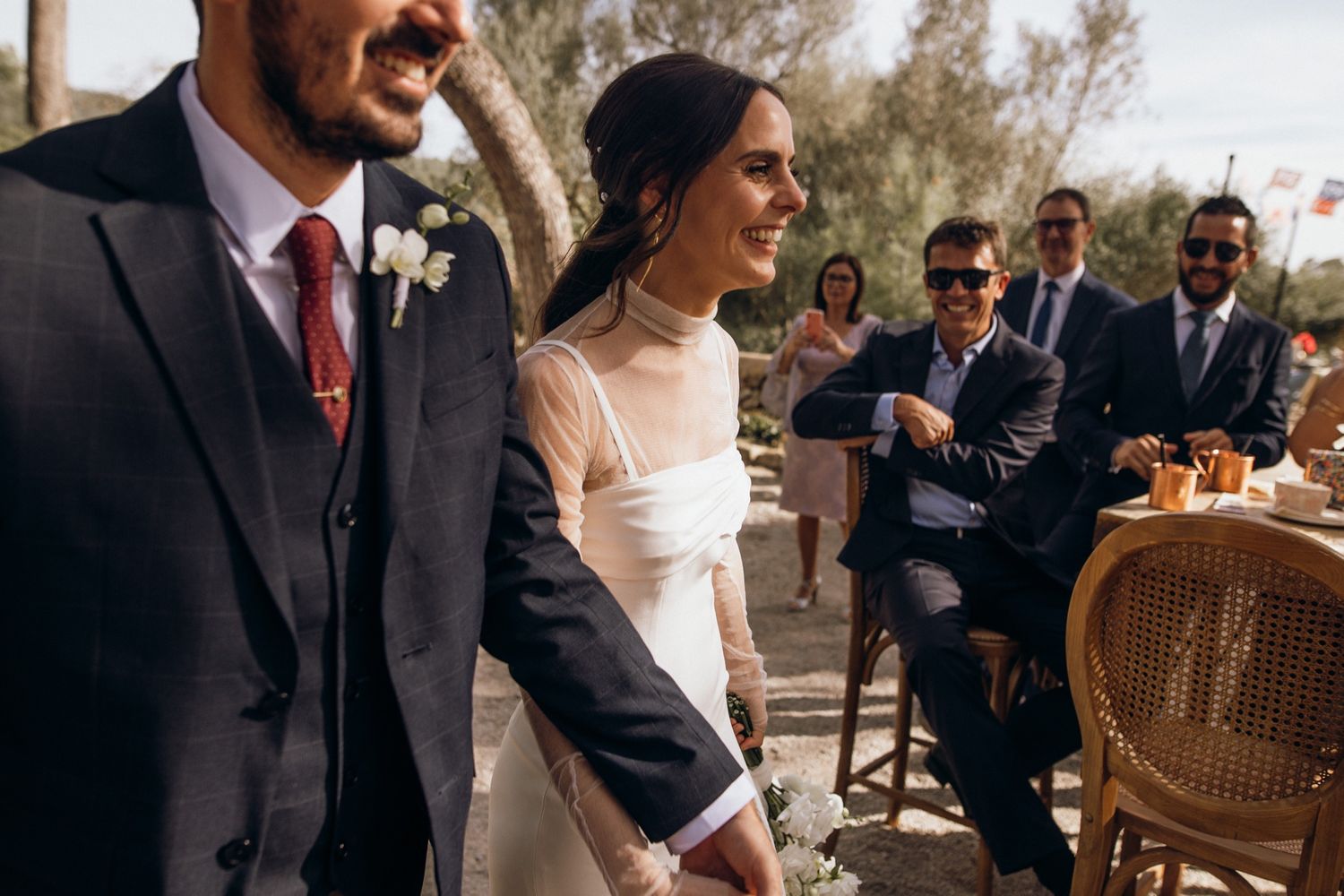 Wedding guests celebrate outdoors at a reception with wicker chairs and natural lighting.