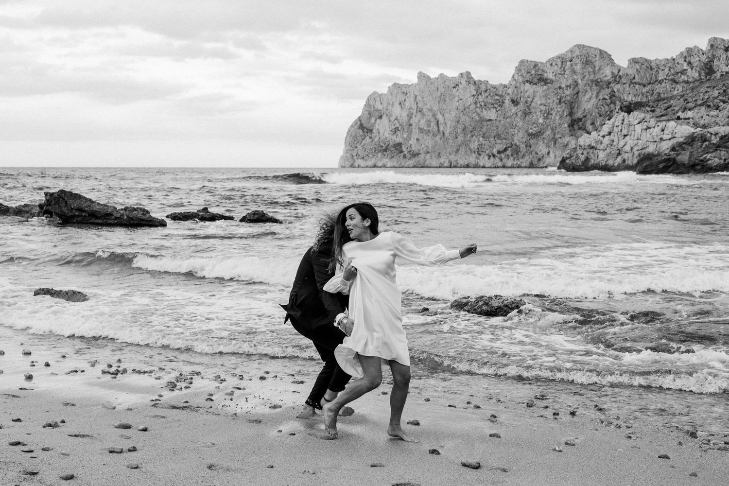 Black and white photo of couple running playfully on rocky beach.
