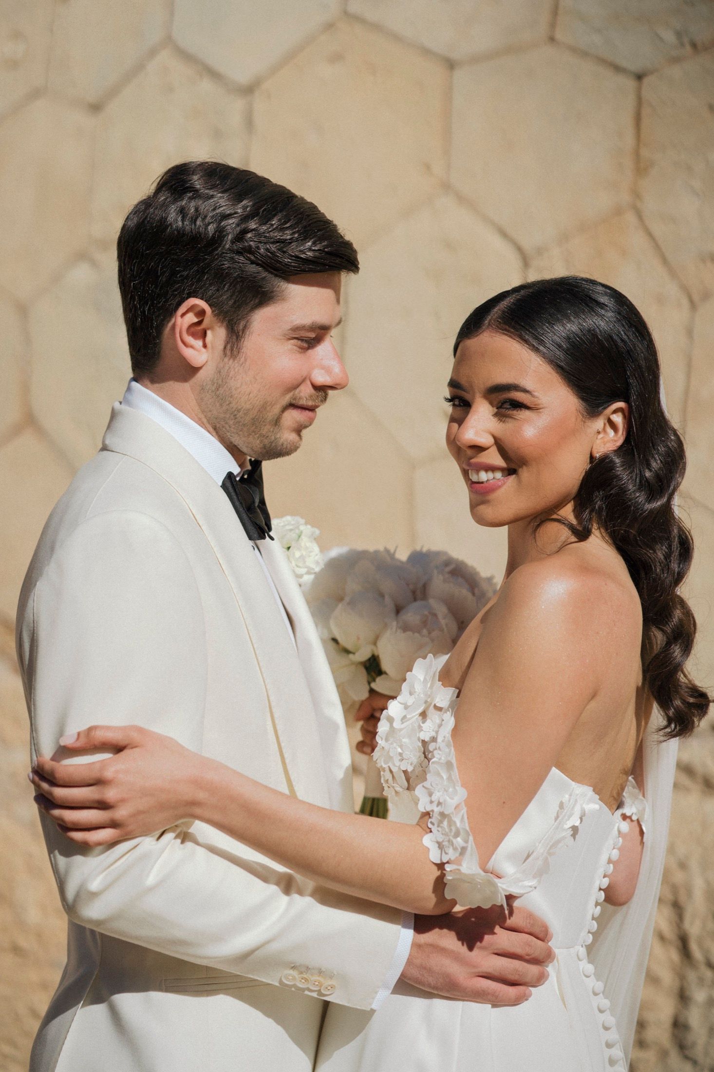 Elegant bride and groom in white attire embrace outdoors, smiling on their wedding day.