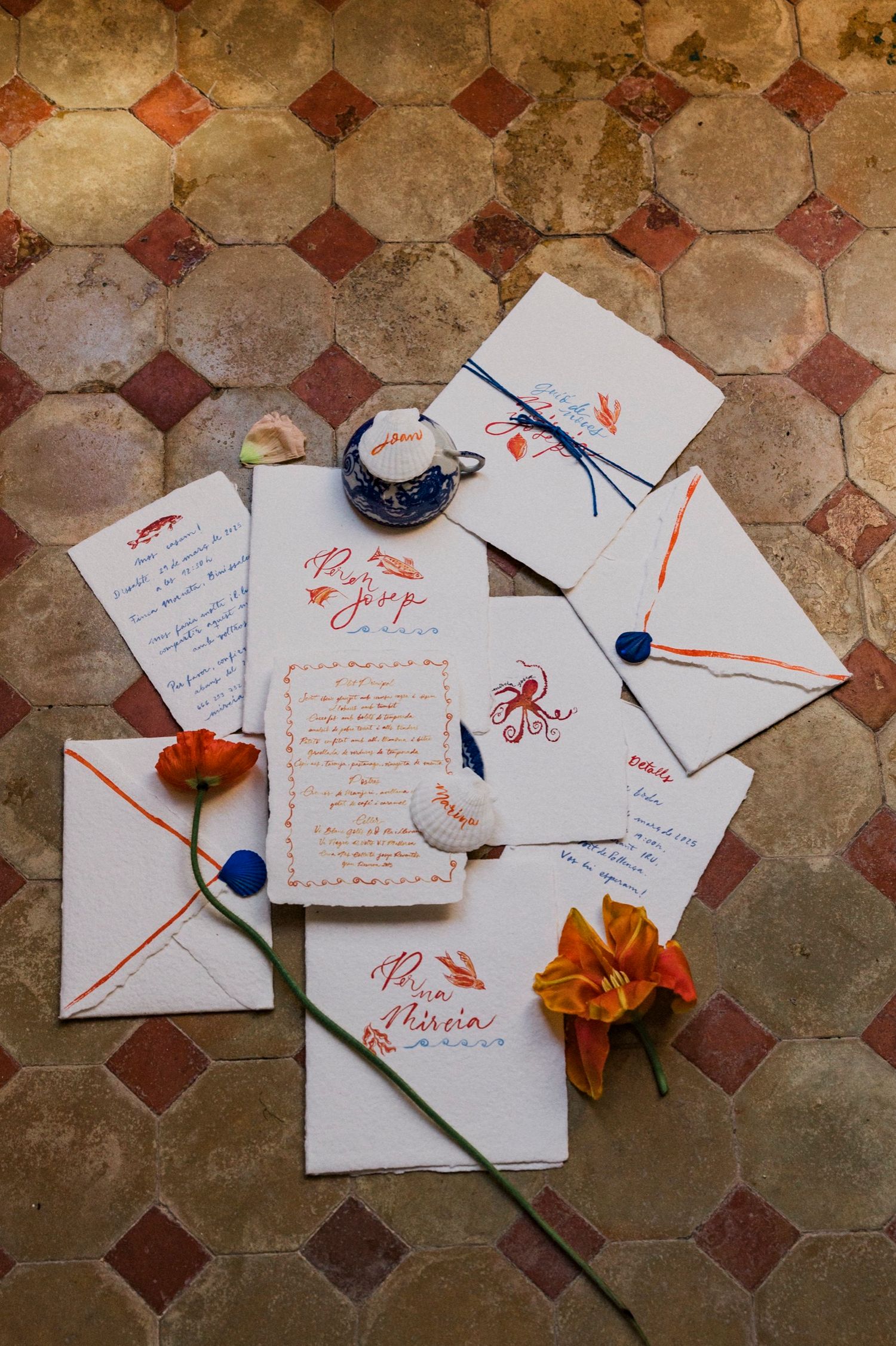 Wedding invitation suite with calligraphy in orange ink, blue wax seals, and orange flowers on a vintage tiled floor.