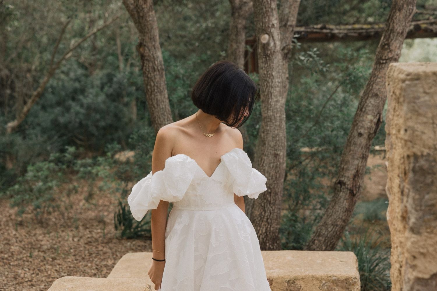 A bride in a white ruffled dress poses in a rustic outdoor setting among trees.