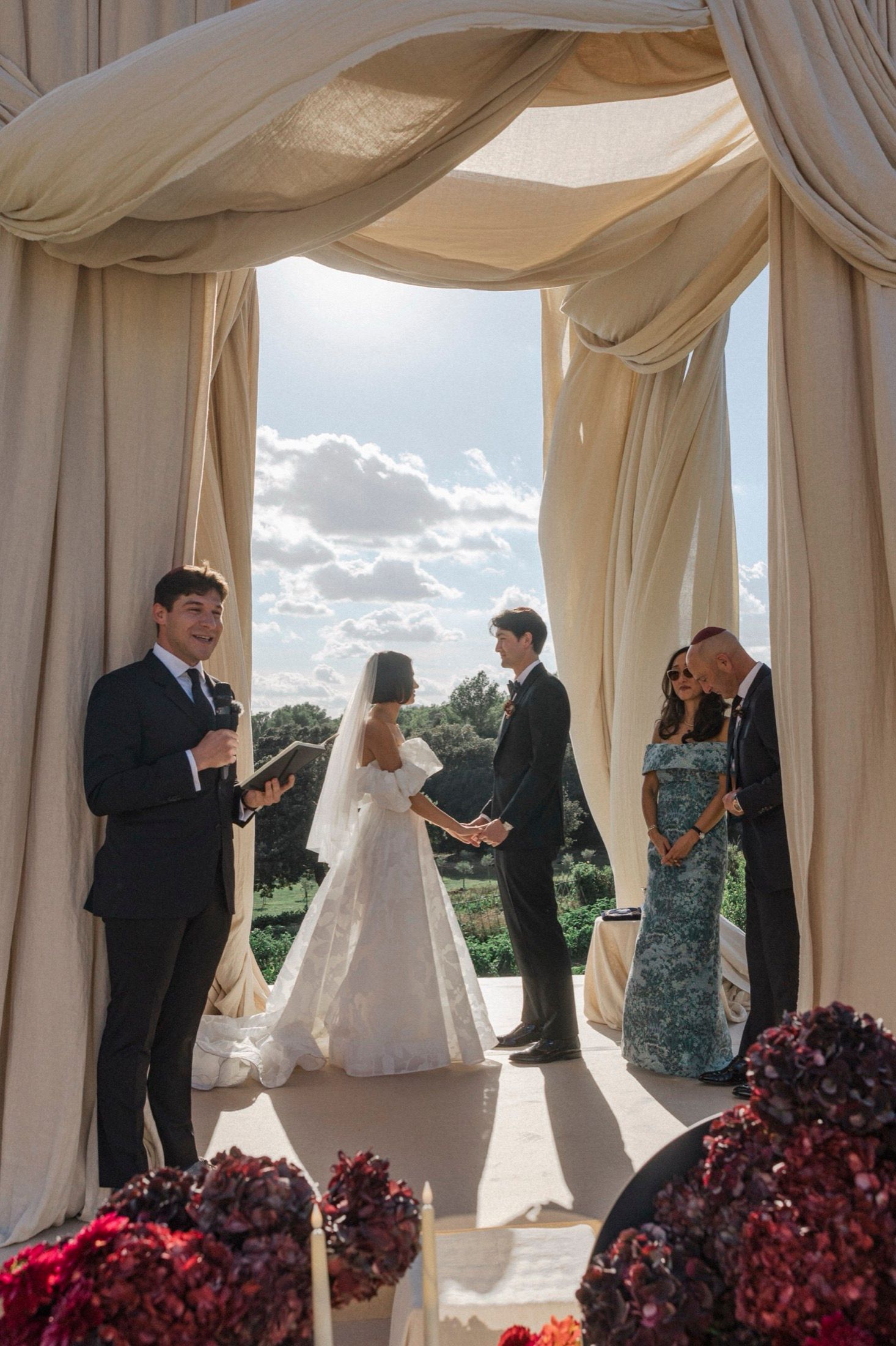 A wedding ceremony takes place under elegant white draped curtains with red flowers in the foreground.