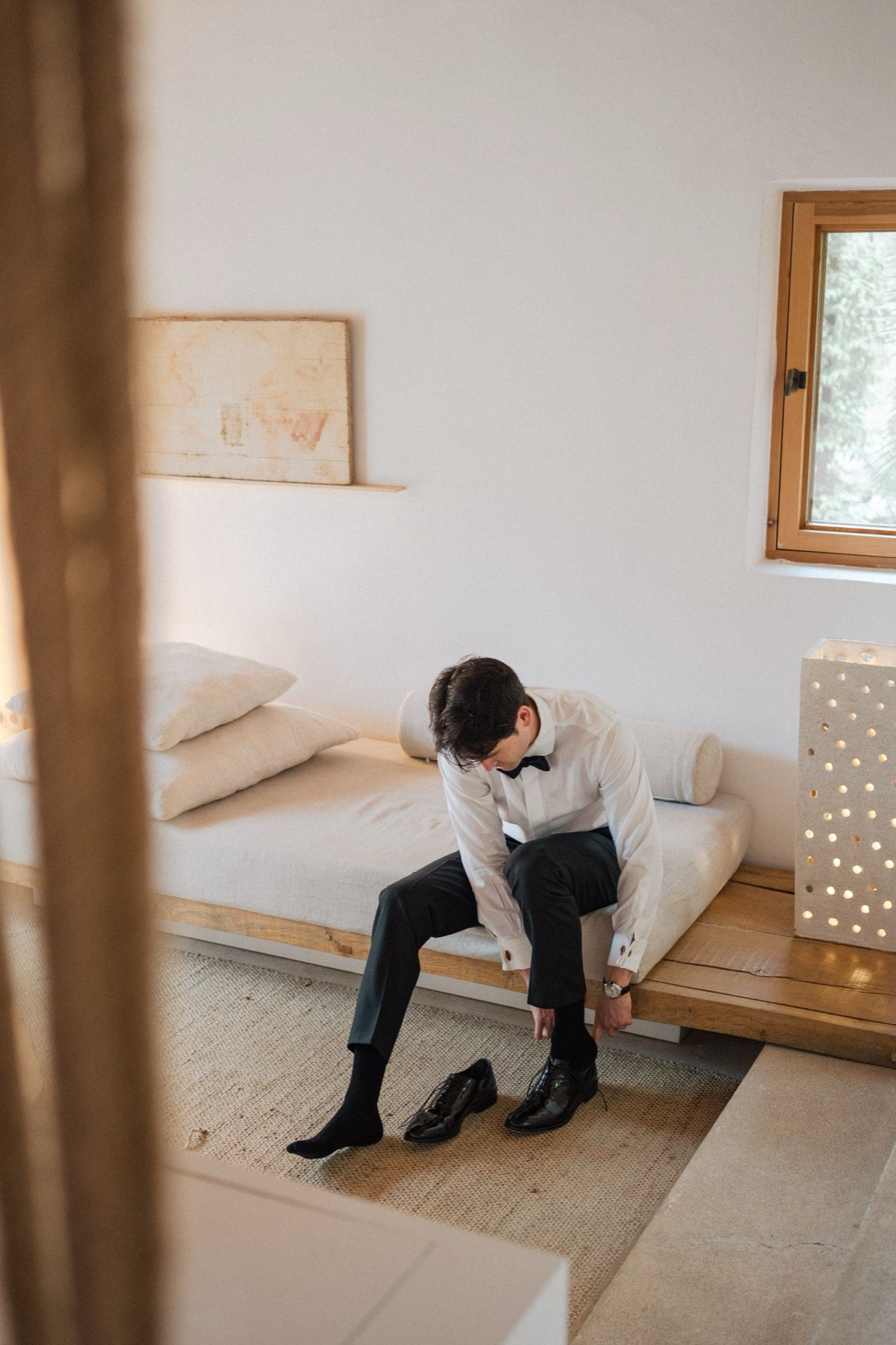 A person in dark clothing sits hunched over on a wooden bench against a white wall with a decorative lamp nearby.