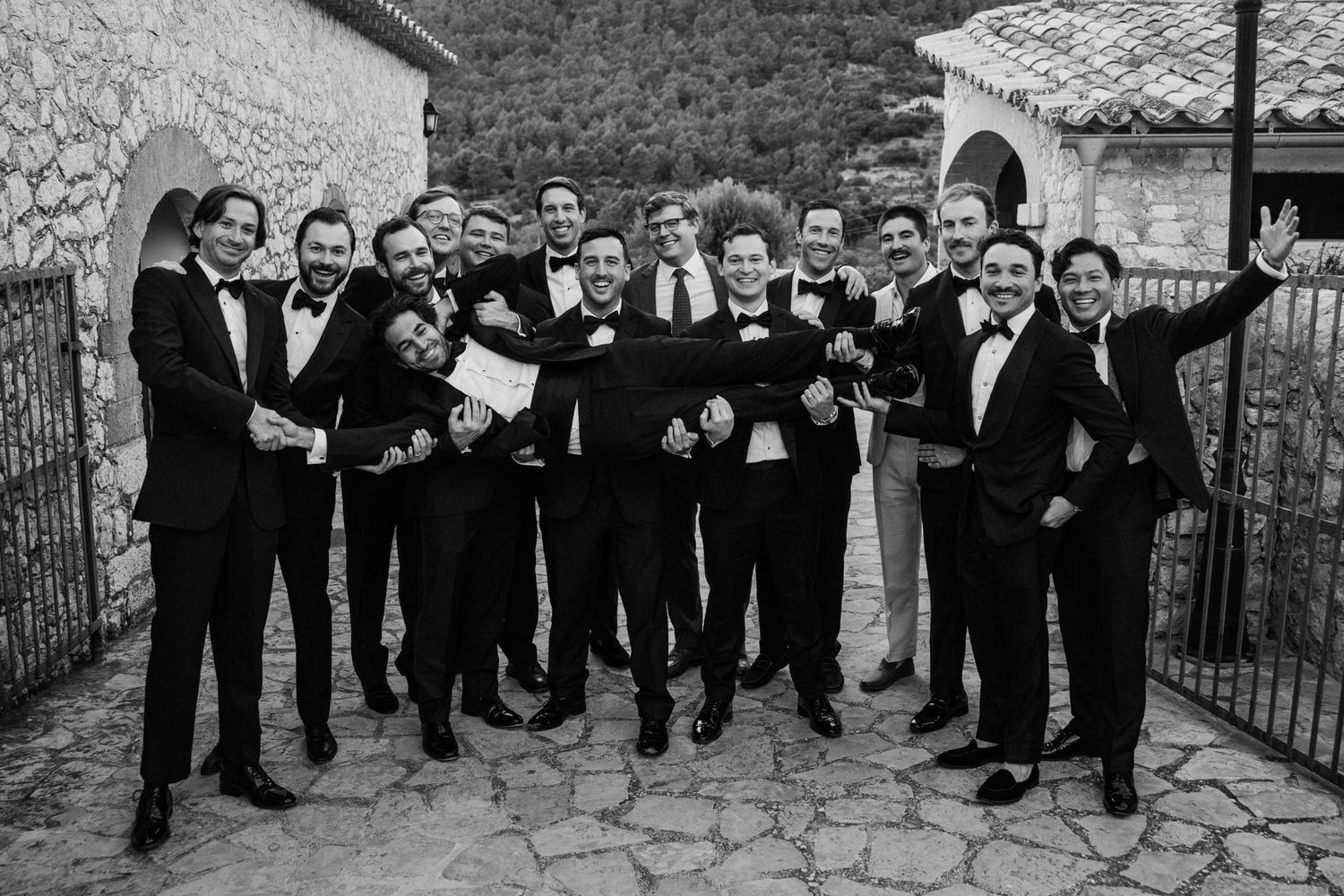 A black and white photo of a wedding party in formal attire gathered outdoors, celebrating joyfully together.