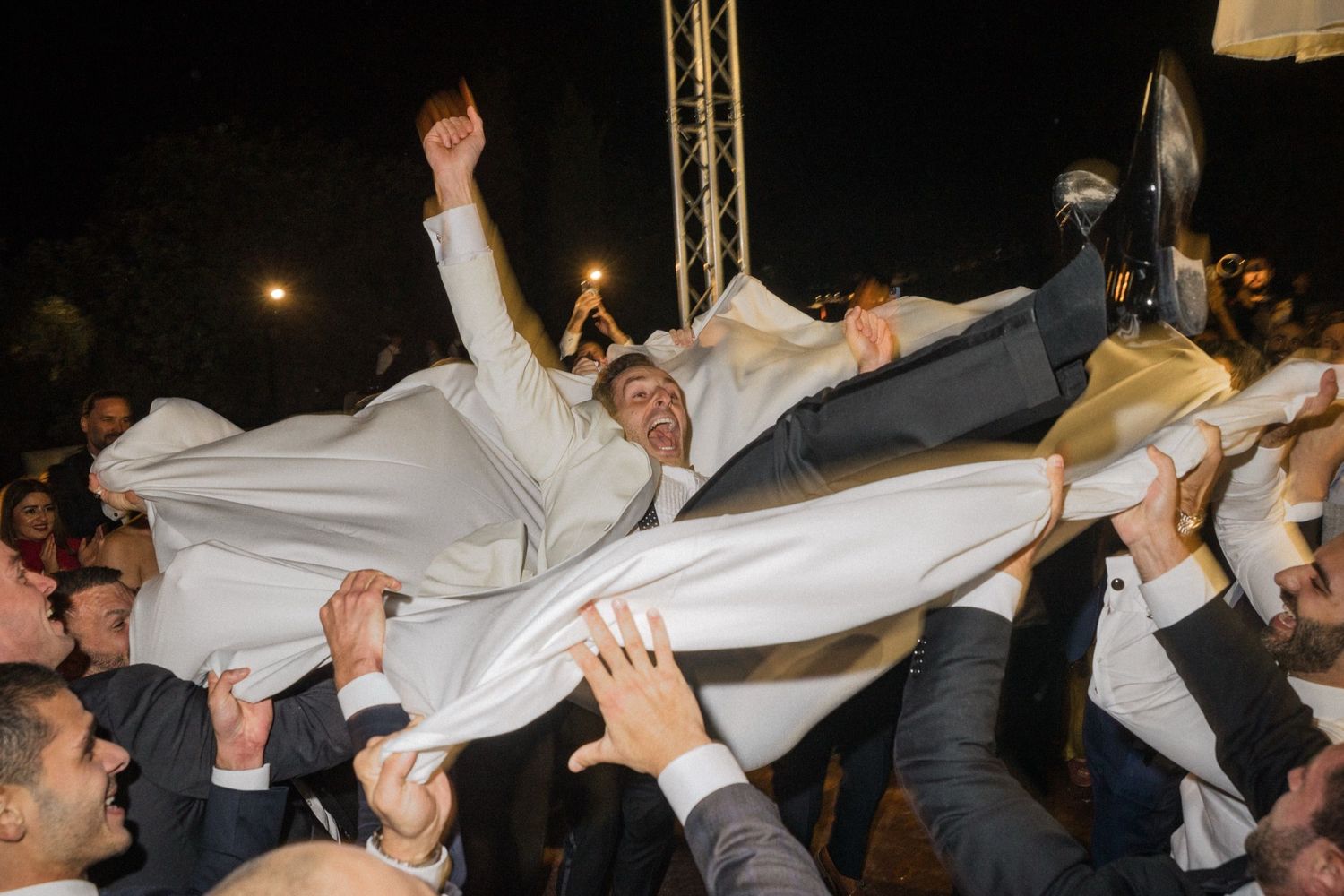 A crowd lifts and tosses someone wearing white formal attire during a celebratory nighttime event.