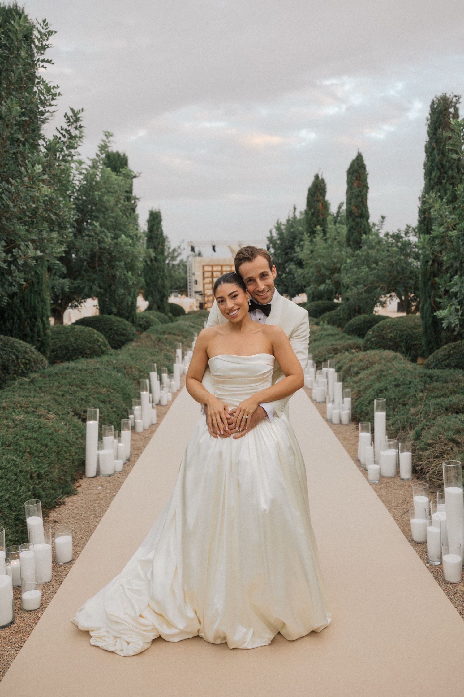 A couple poses on an elegant garden path lined with glowing candles and tall cypress trees at dusk.