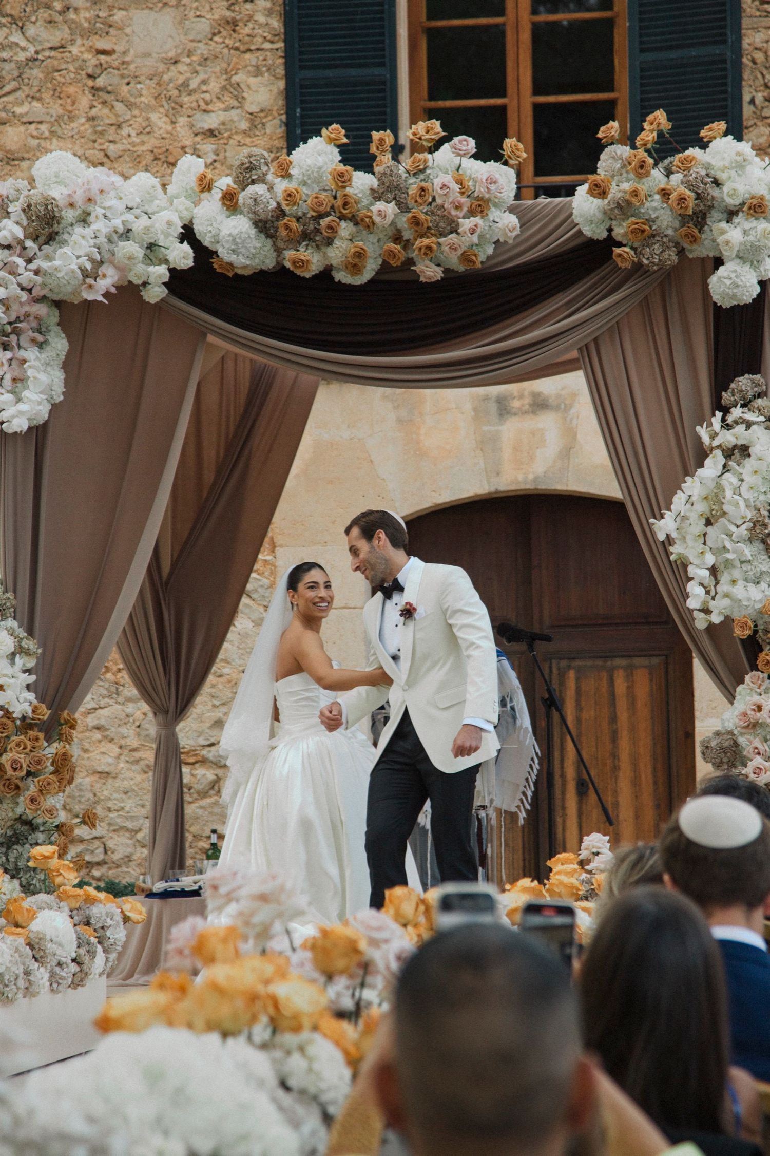 Elegant wedding ceremony setup with brown draping and white and peach floral arch decorations at a stone venue.