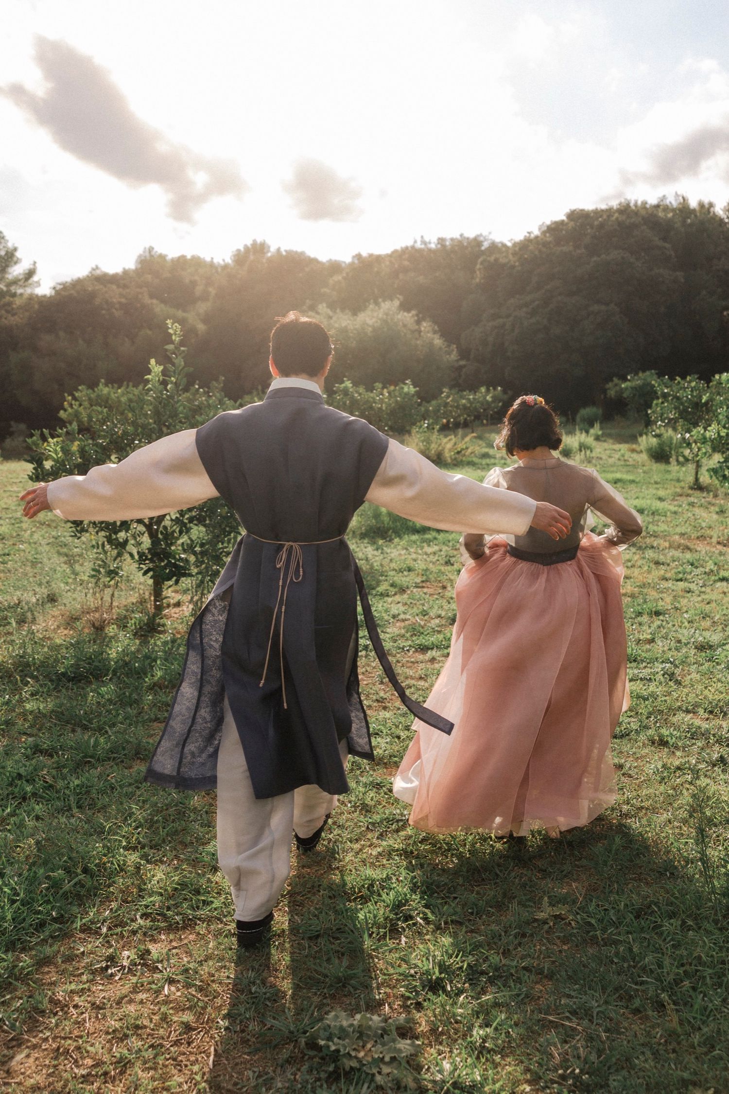 Three people with arms outstretched walking through a sunlit field at sunset.
