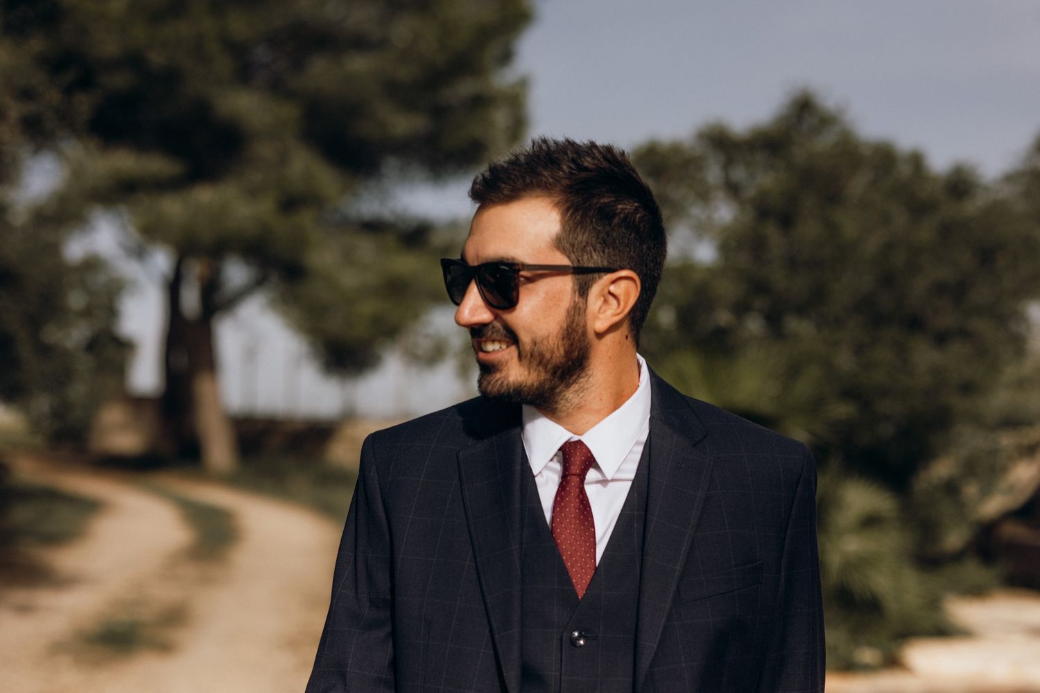 Professional wearing a dark suit and red tie stands in an outdoor setting with natural lighting.