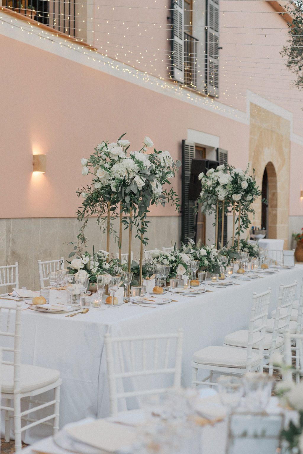 Elegant white wedding reception tables set against a pink building with string lights overhead.