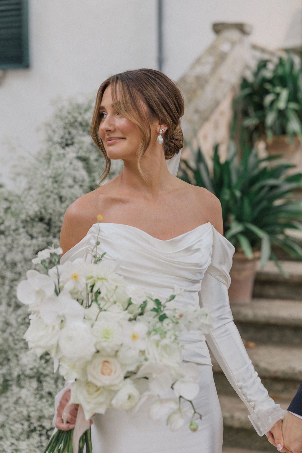 A bride in an elegant off-shoulder white gown holds a bouquet of white flowers while smiling.