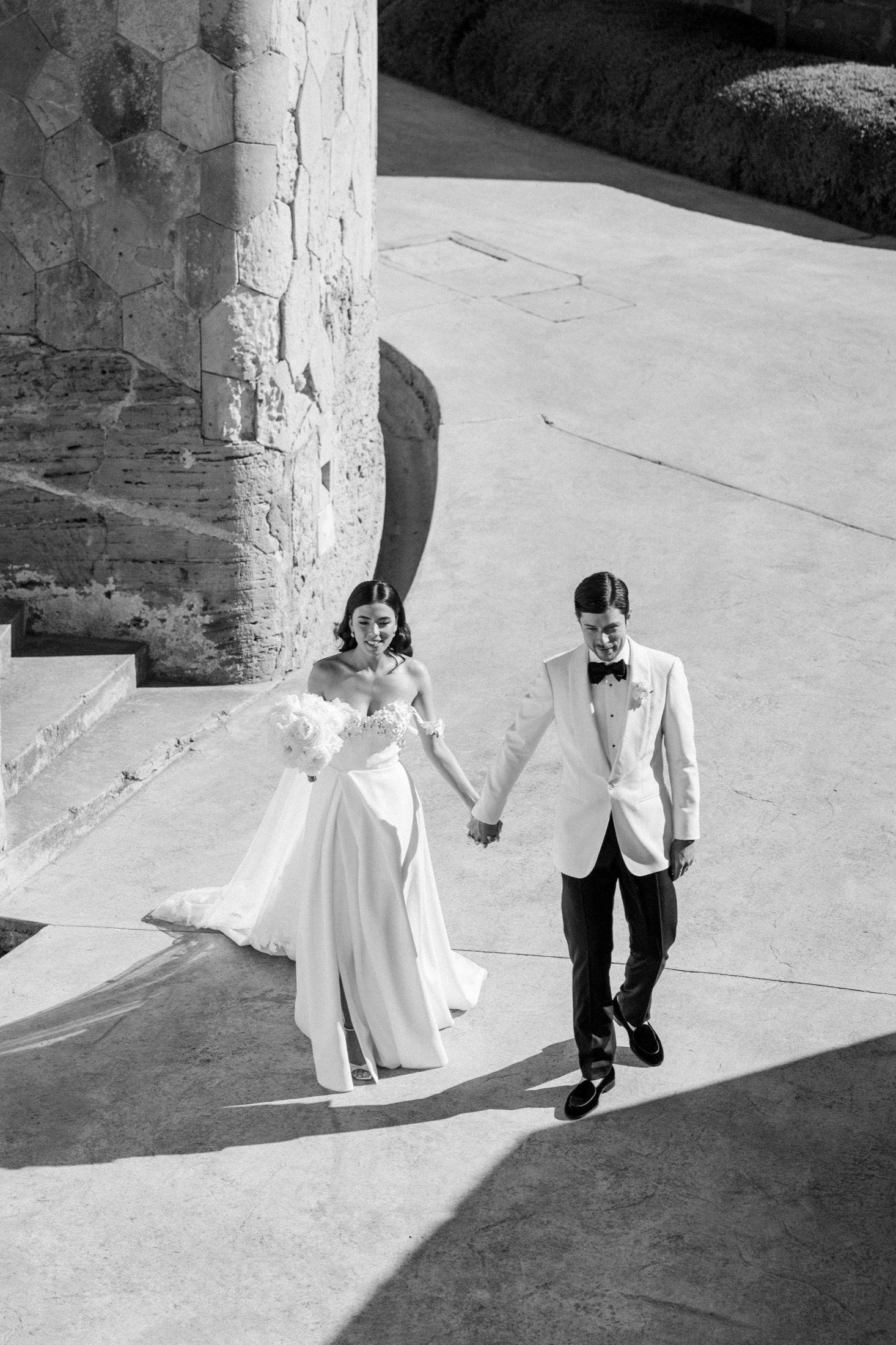 Black and white photo of bride and groom holding hands, walking near stone columns on their wedding day.
