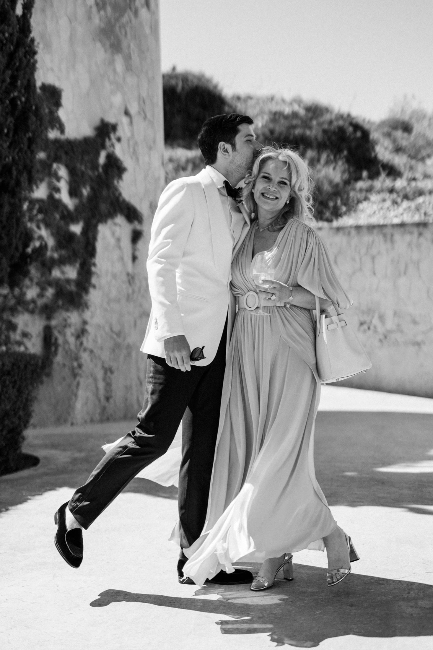 Black and white photo of a groom kissing his smiling bride on the cheek as they walk together outdoors.
