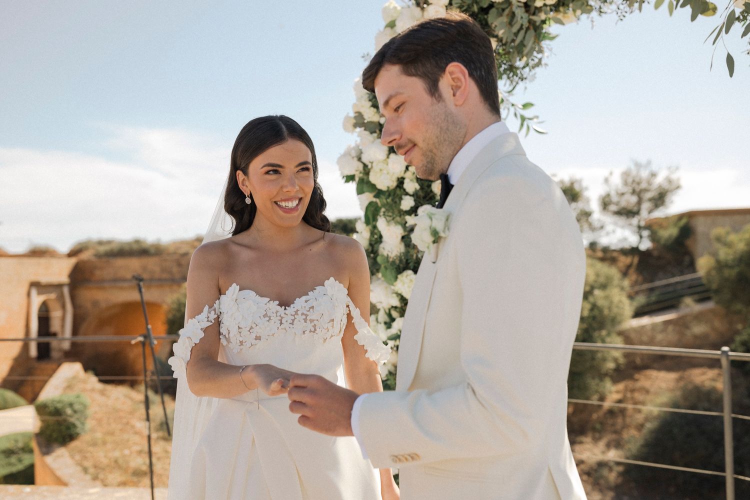 Couple exchanging vows at outdoor wedding ceremony with floral arch, bride in lace gown and groom in white suit.