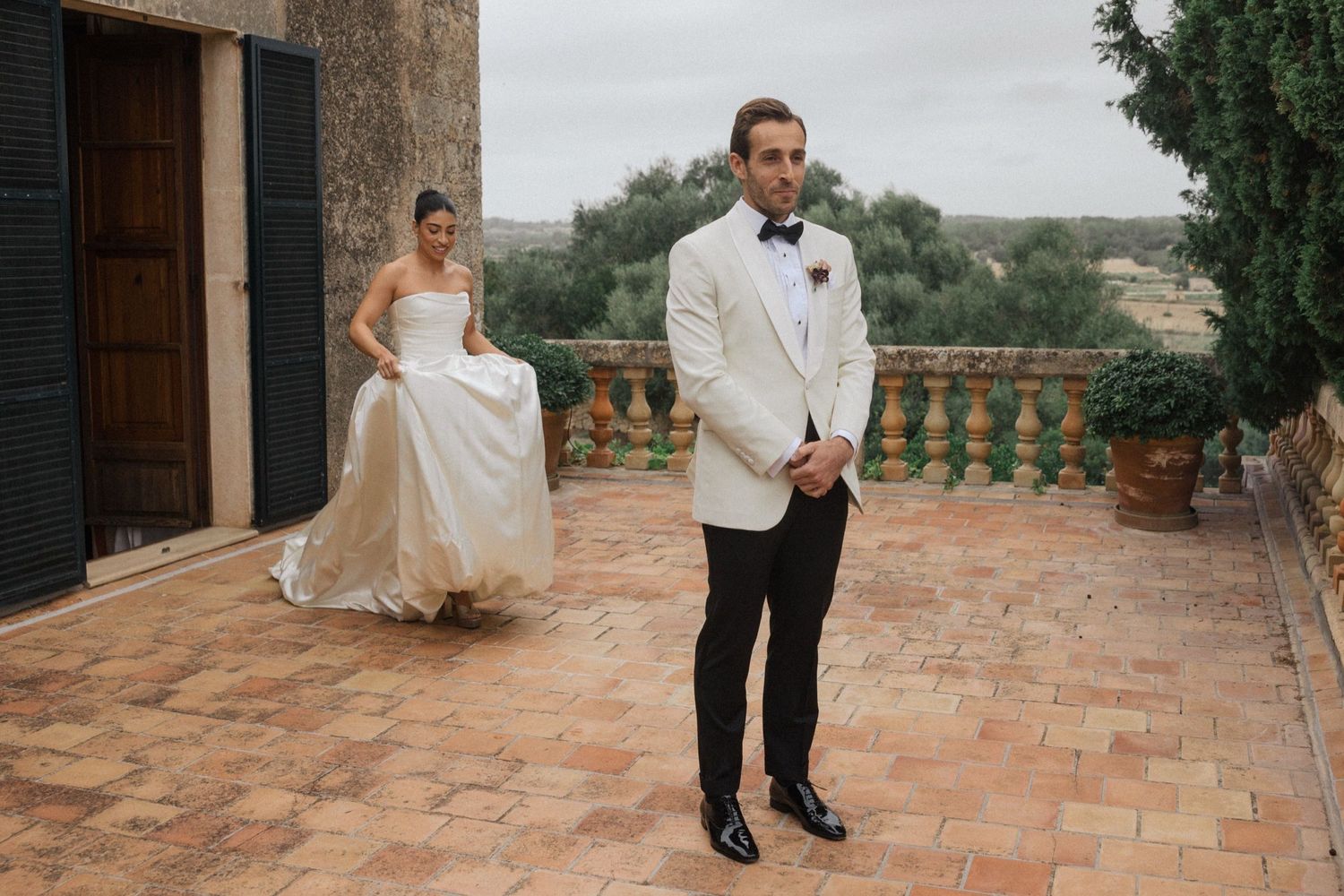 A couple in elegant wedding attire poses together on a rustic terrace with stone balustrades and scenic views.