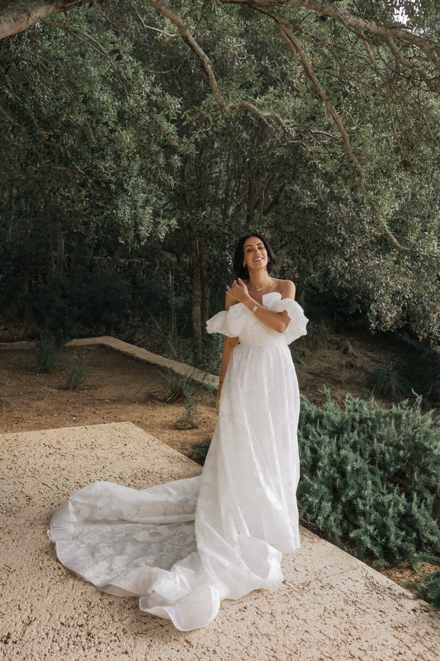 A person in a flowing white wedding dress twirls gracefully in a natural outdoor garden setting.