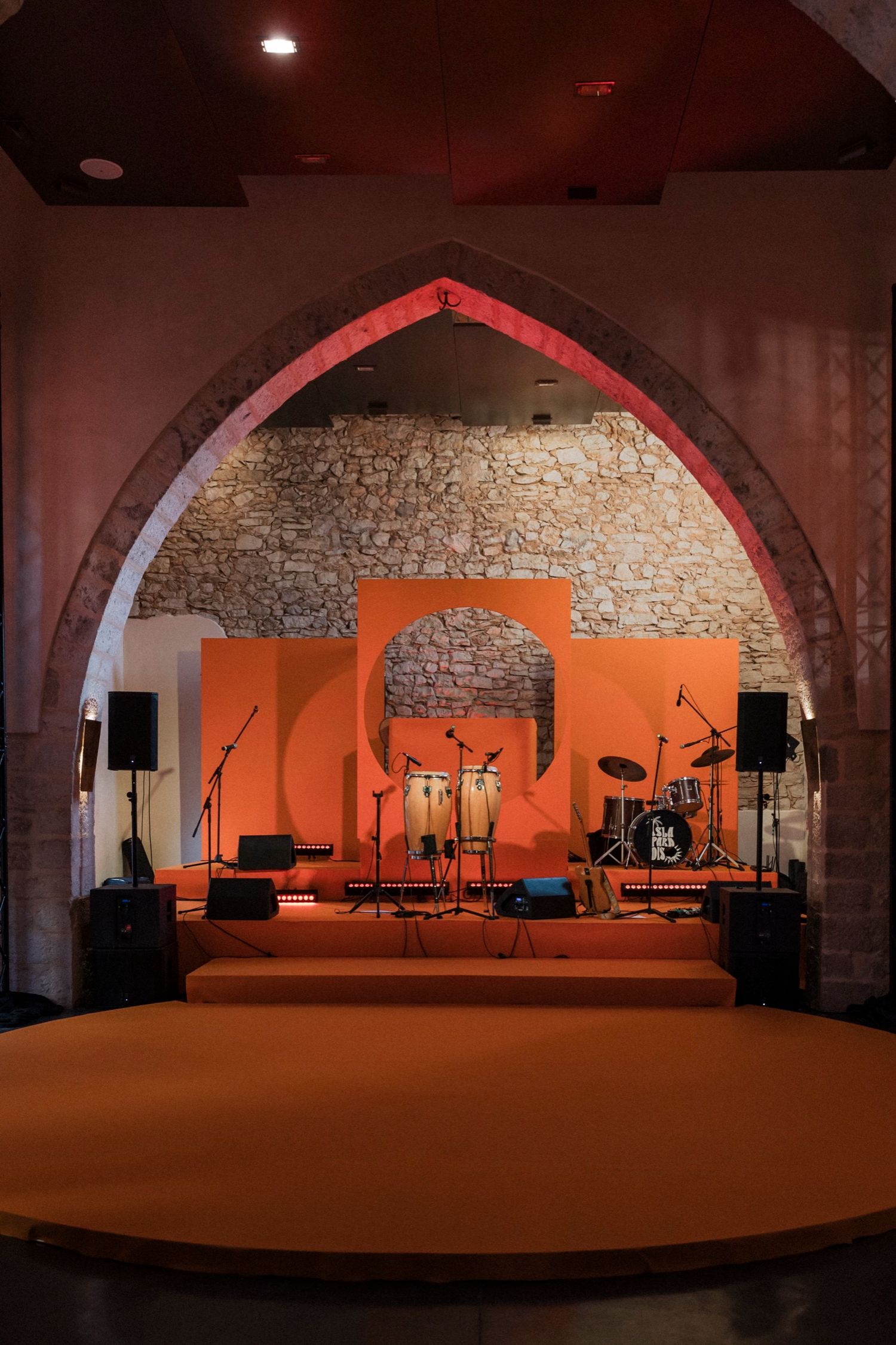 Empty stage setup with orange walls and stone archways in a rustic performance venue illuminated by warm lighting.