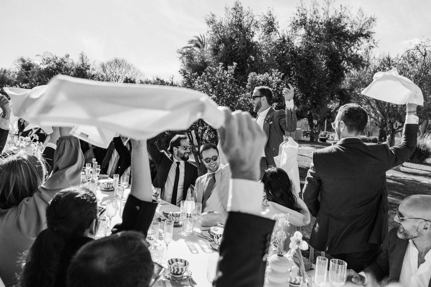 Wedding guests holding white umbrellas gather outdoors during a sunny garden ceremony.