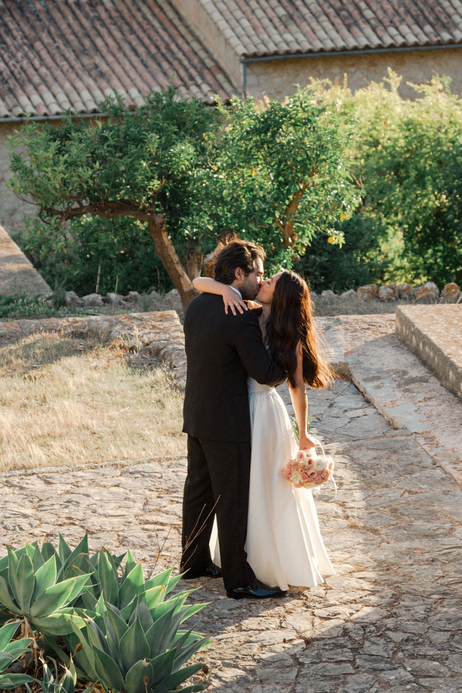 A couple embraces on a stone path surrounded by lush greenery and agave plants during a romantic outdoor moment.