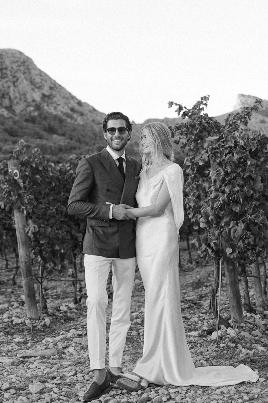 A couple poses together in a scenic vineyard setting with mountains in the background during a wedding photoshoot.