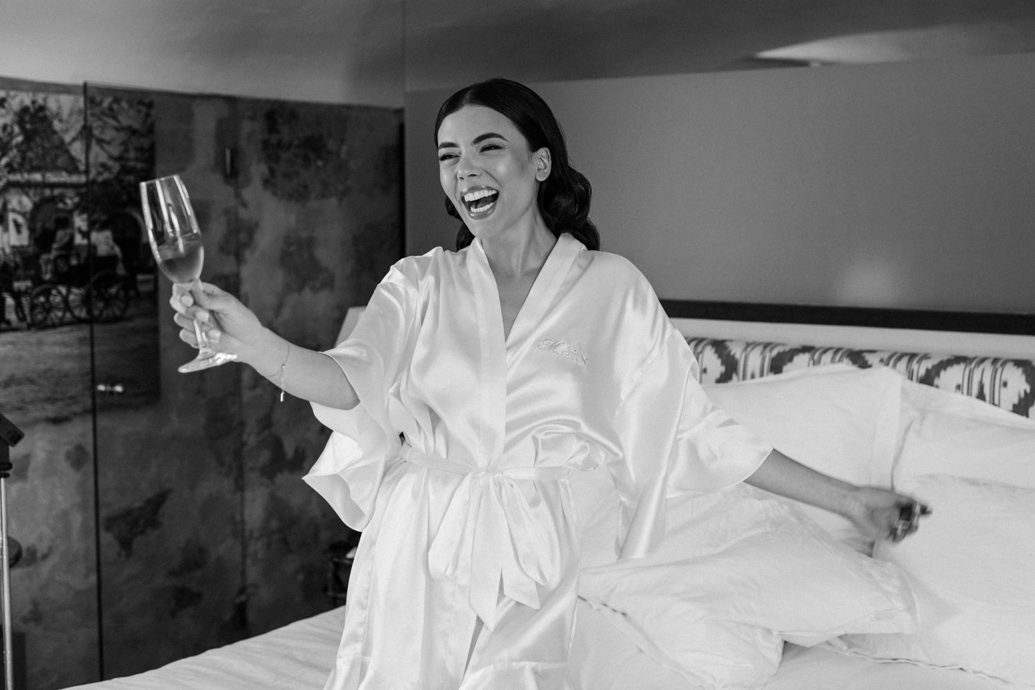 Woman in white satin robe laughing and raising a wine glass while sitting on a bed, black and white photo.