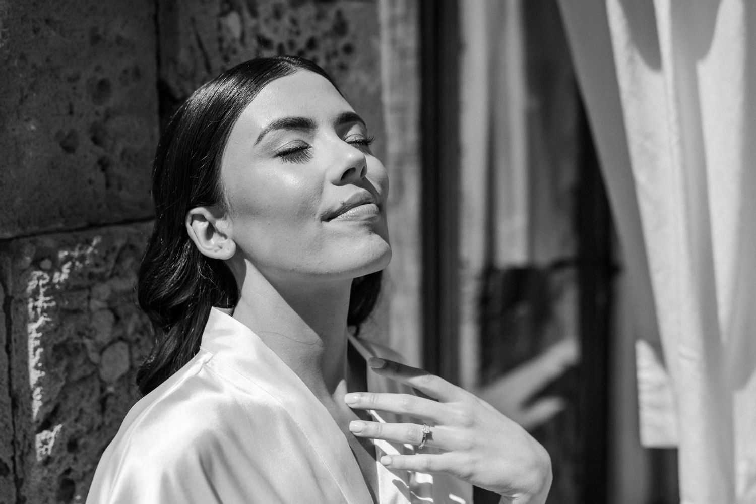 Black and white portrait of a serene woman in a silk robe, eyes closed, enjoying sunlight outdoors.