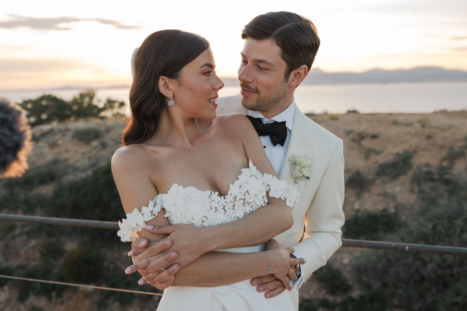 Couple in wedding attire embracing at sunset, groom in white tuxedo, bride in floral off-shoulder gown.