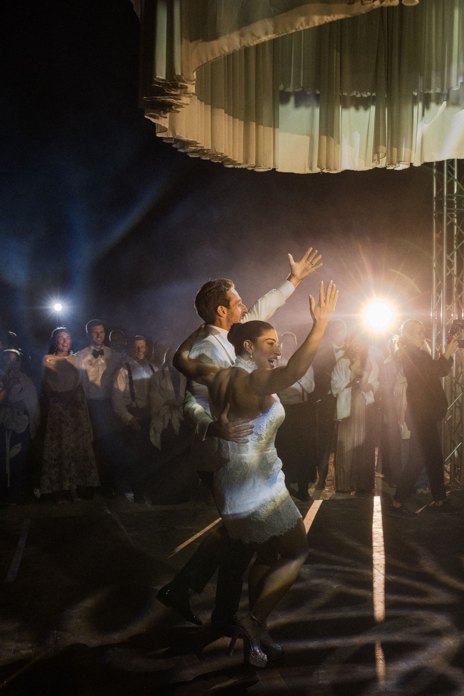 Dancers performing a dramatic sequence in low lighting with bright spotlights creating silhouettes at a wedding reception.