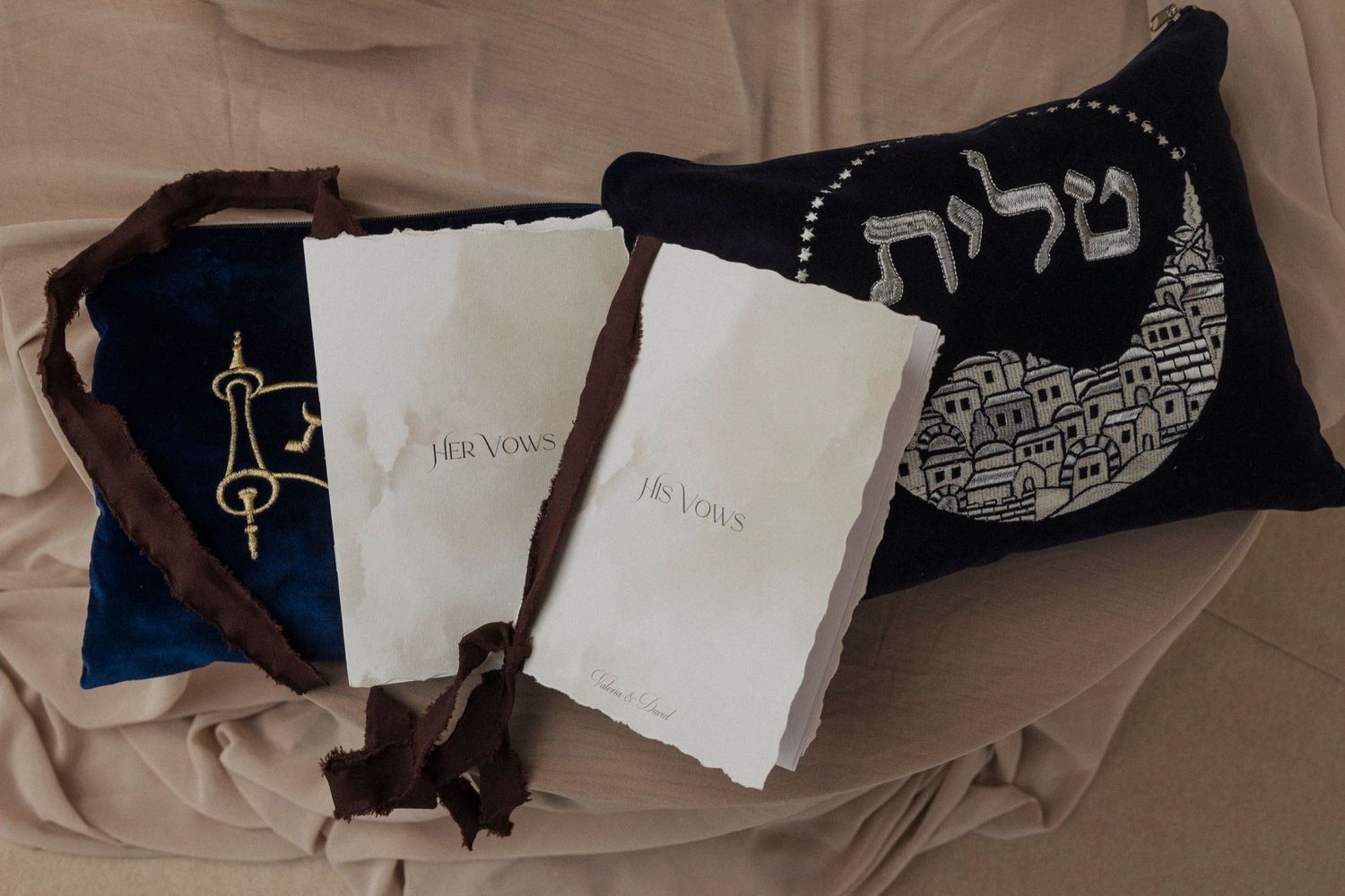 Jewish prayer items including white prayer books and embroidered tallit bags arranged on beige fabric.