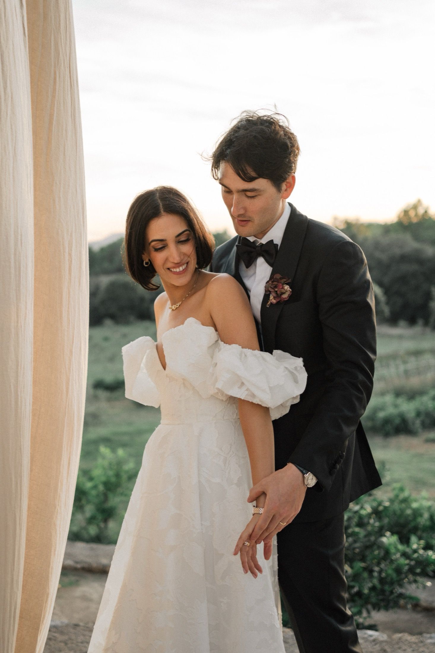 A romantic wedding couple embraces outdoors, with the bride in an off-shoulder white gown and groom in classic tuxedo.