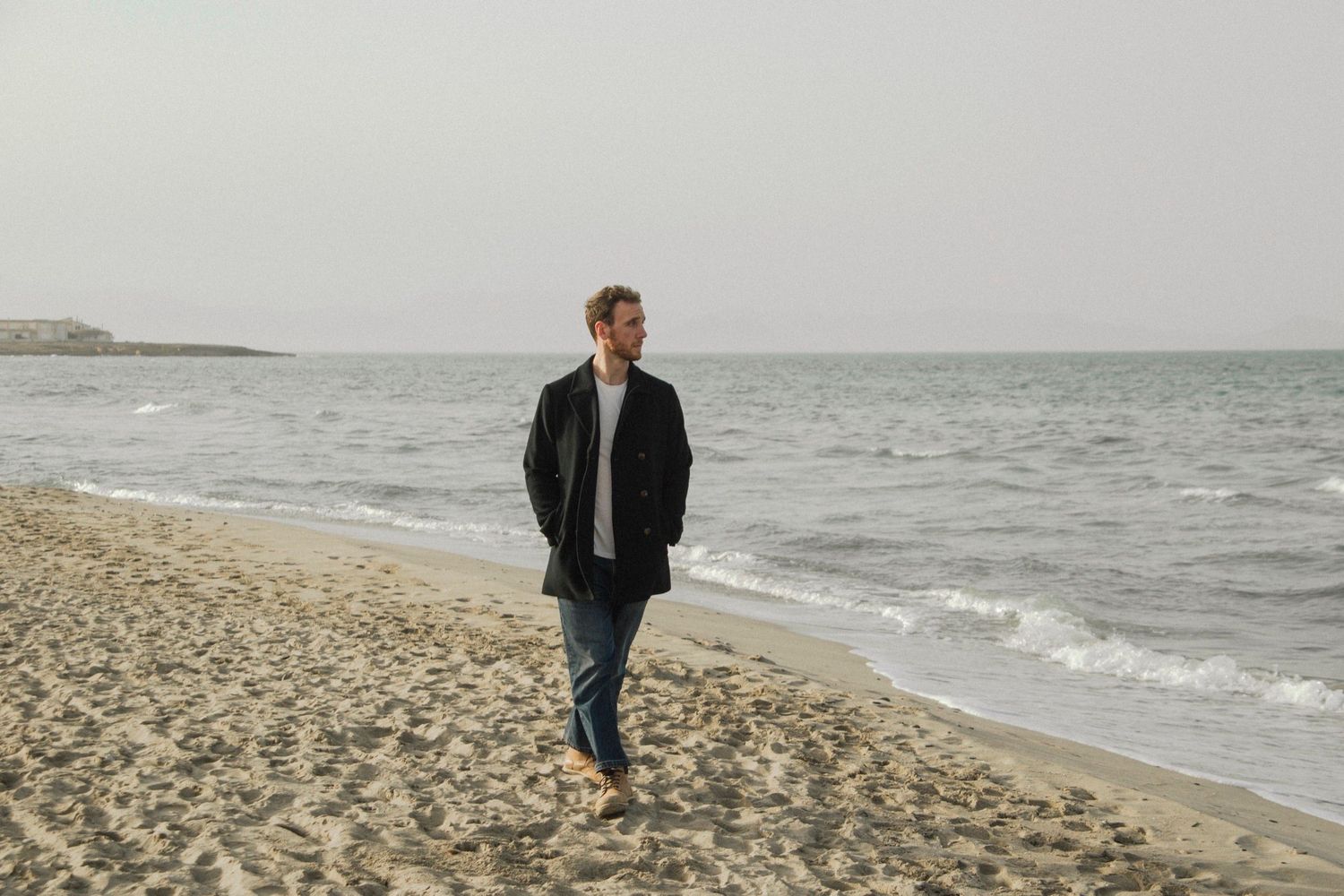 A person walks along a misty beach shoreline with waves crashing against the sand on an overcast day.