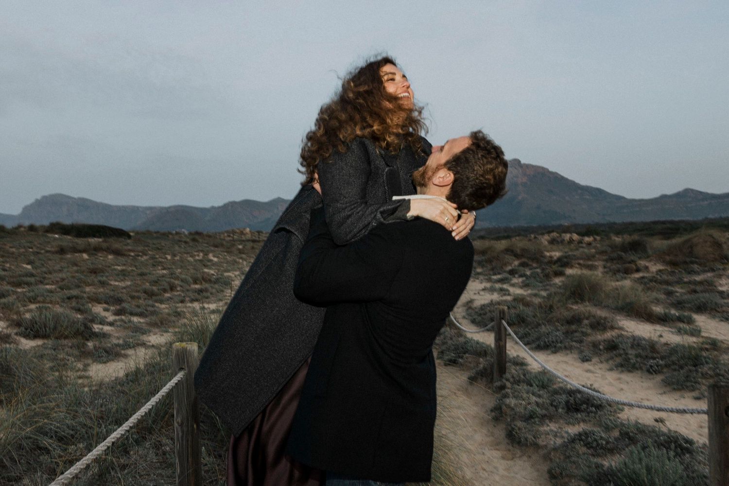 Couple embraces in dramatic black attire against desert landscape at sunset creating silhouette effect.