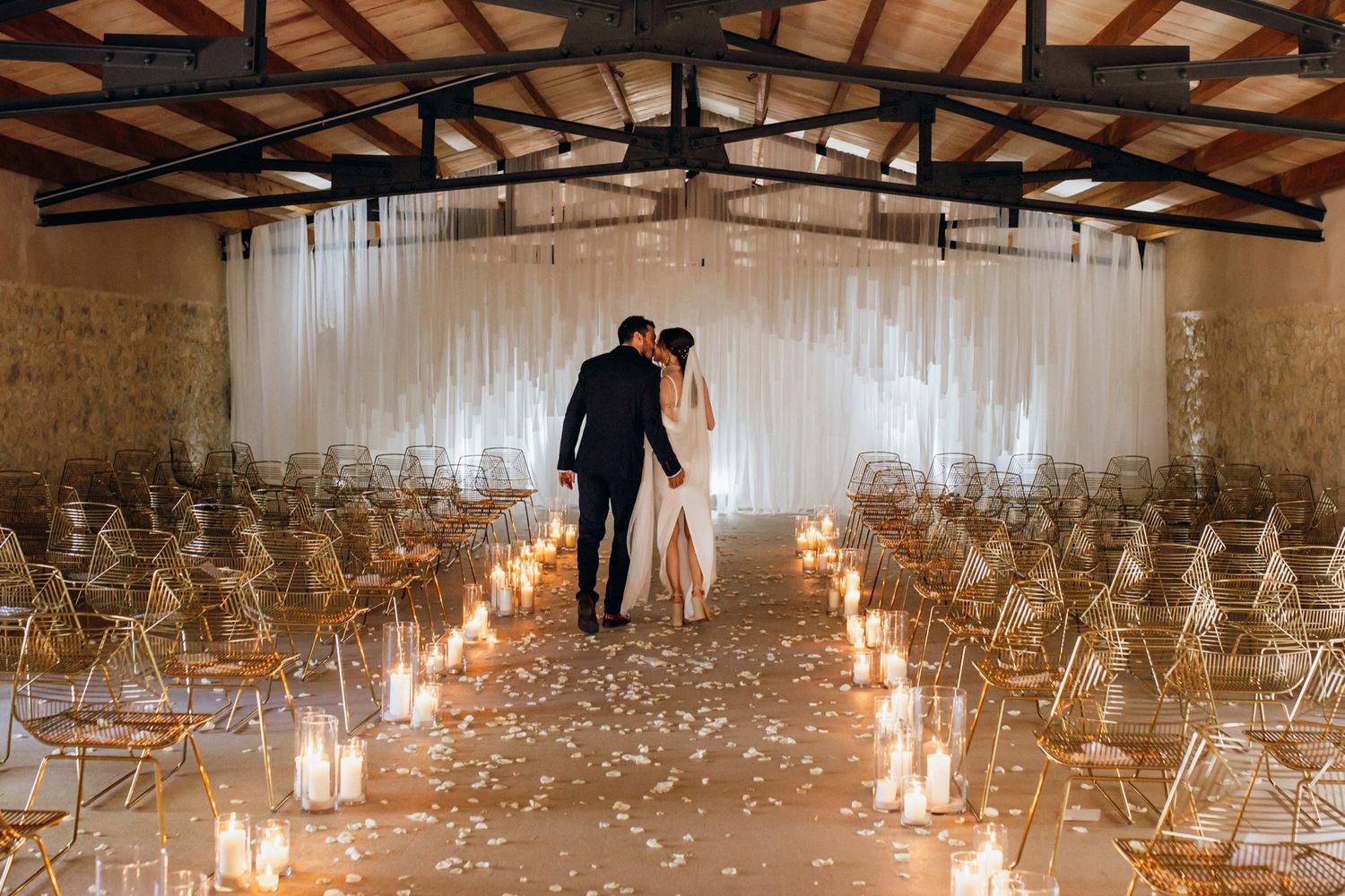 Rustic wedding ceremony setup with wooden beams, rows of chairs, and romantic lantern lighting along the aisle.