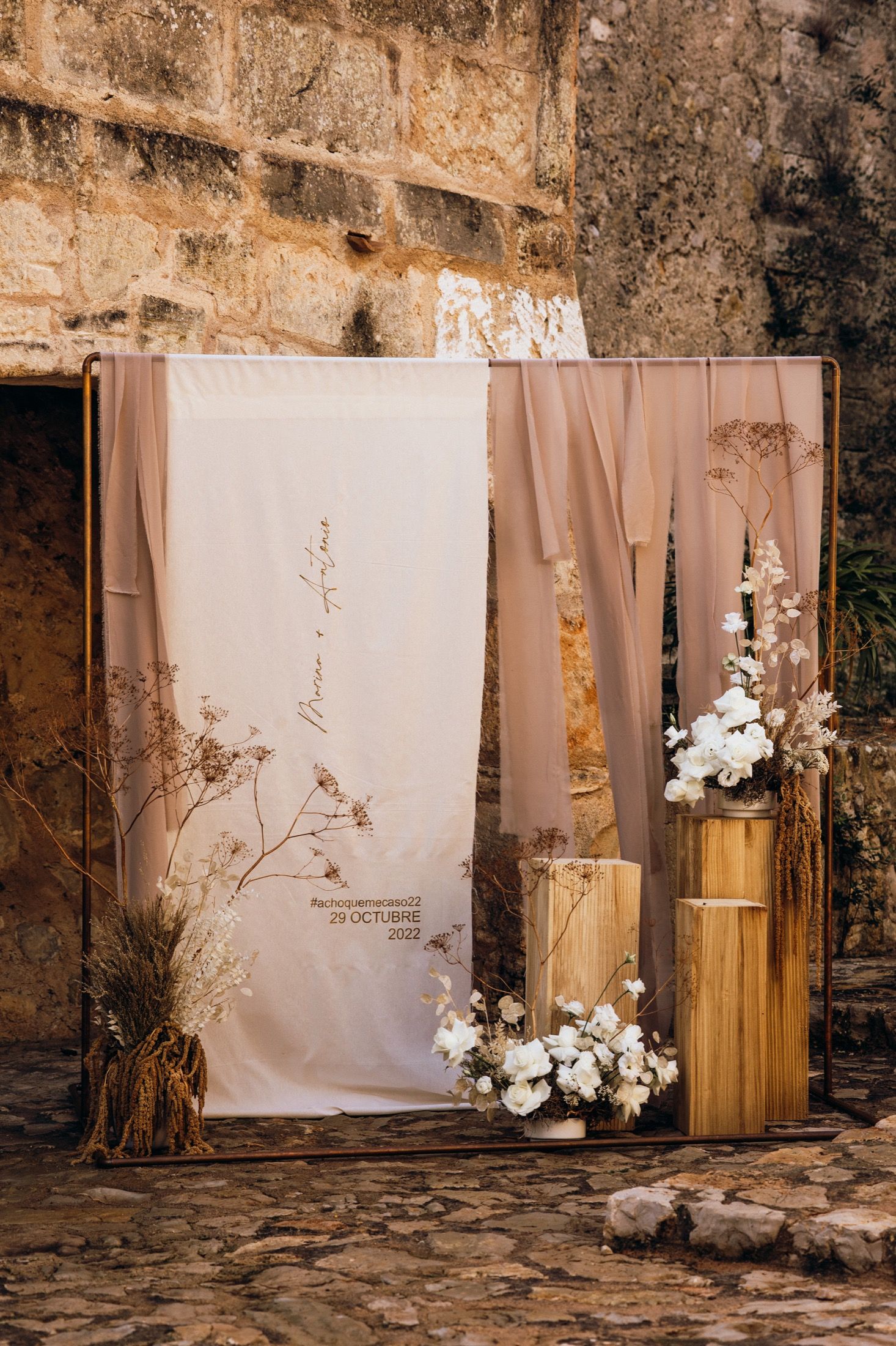 Rustic wedding photo backdrop with white curtains, wooden pedestals, and floral arrangements against a weathered stone wall.