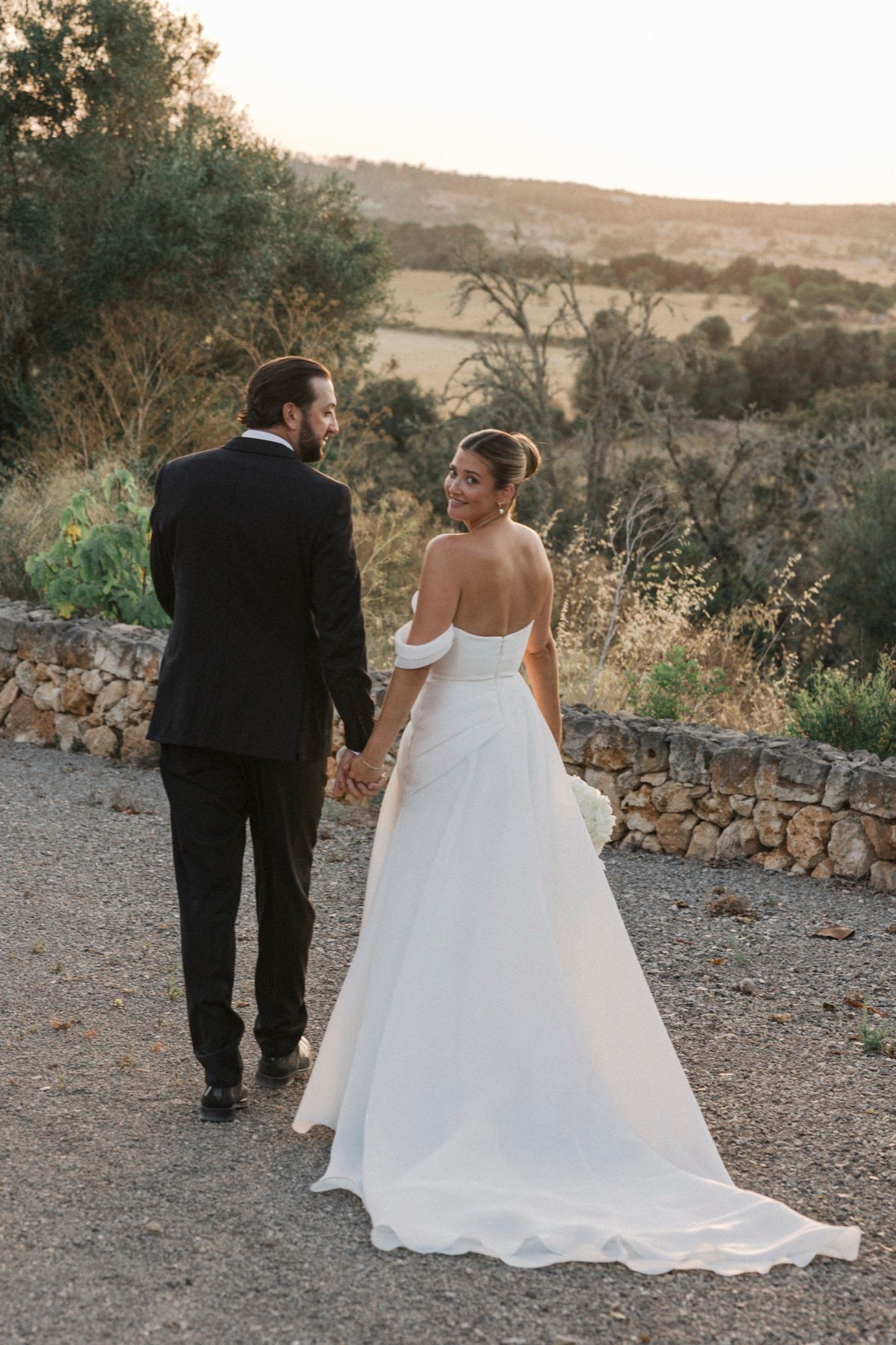 Newlyweds walk together along a gravel path with stone walls and rolling hills in the background at sunset.