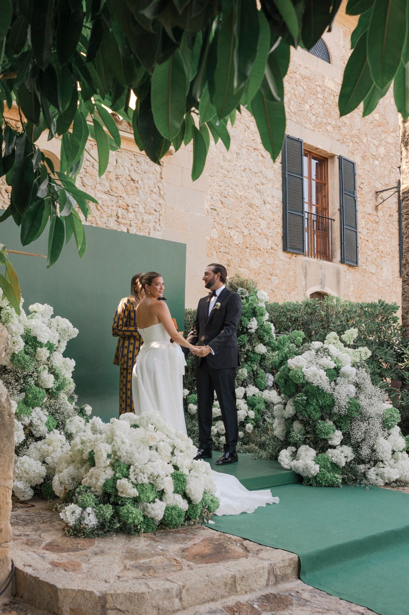 Outdoor wedding ceremony setup with lush white hydrangea arrangements against a stone wall backdrop.