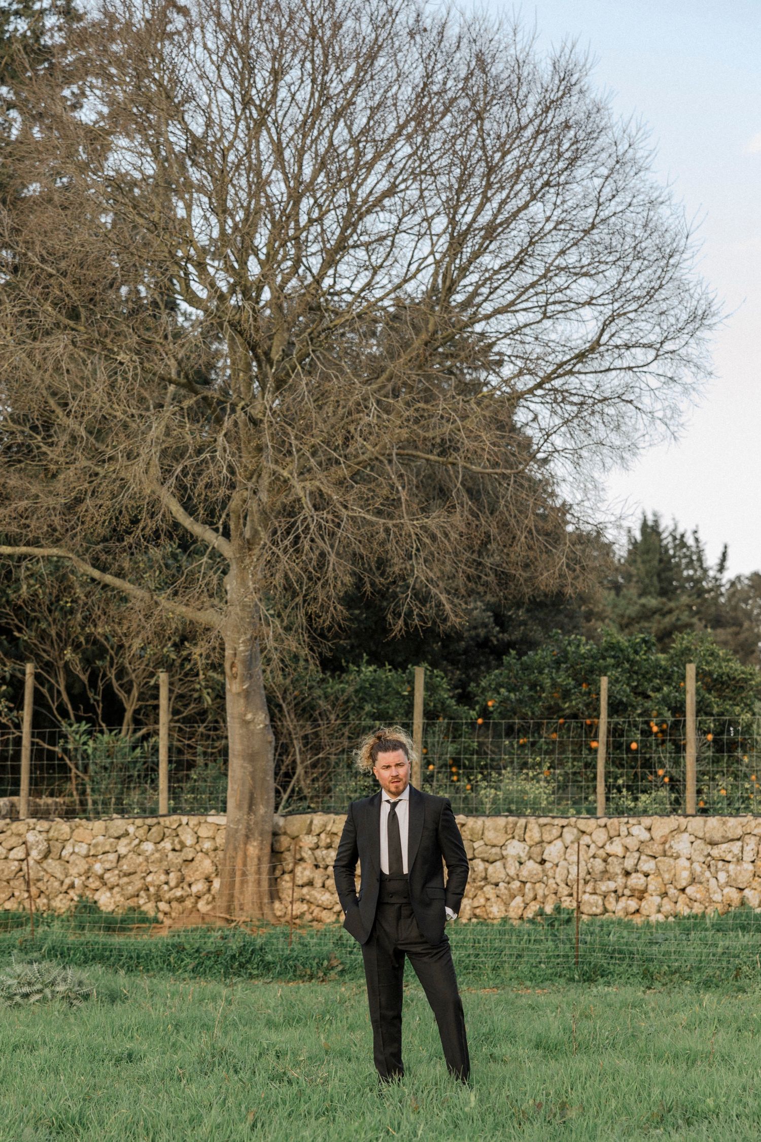 Figure in formal attire poses near stone wall and bare trees in countryside setting.