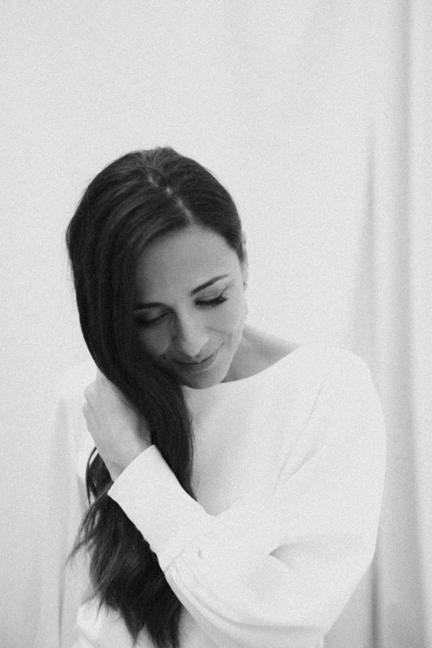 A candid black and white portrait of someone in a white sweater looking down with a gentle smile.
