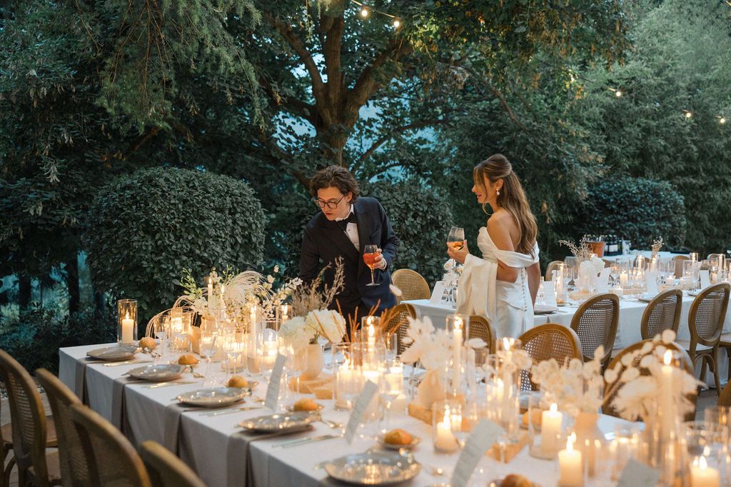 Candlelit wedding reception tables decorated with white linens and rustic chairs under string lights.