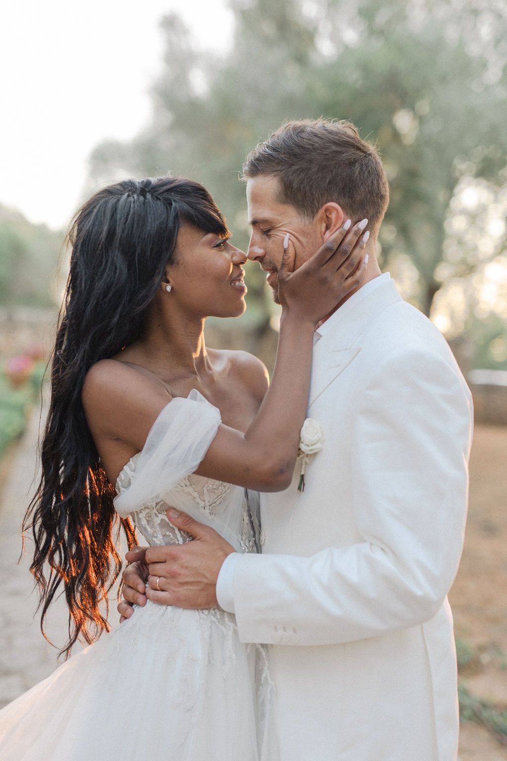 Newlyweds embracing in white attire against rustic Mediterranean backdrop.
