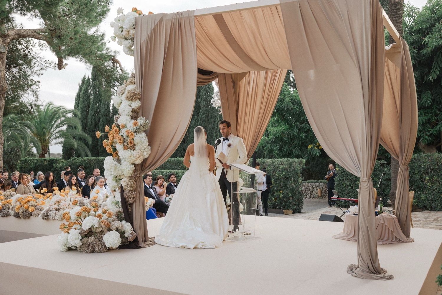 Elegant outdoor wedding ceremony setup with flowing beige drapery and natural greenery backdrop.