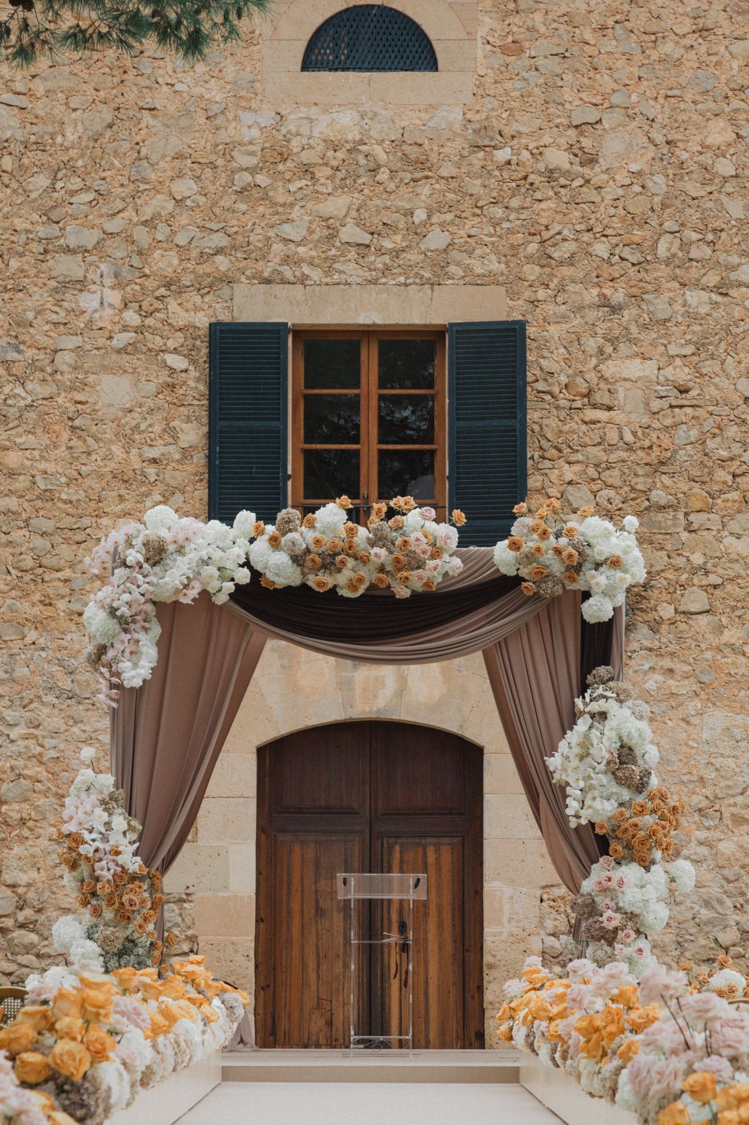 Elegant wedding arch with white and peach floral arrangements against a rustic stone wall with wooden shuttered windows.
