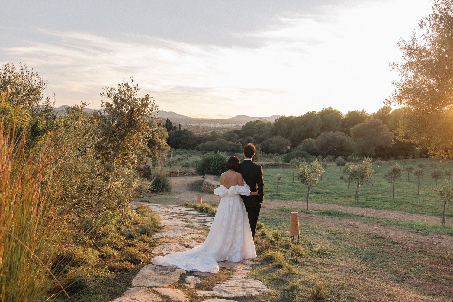 Couple in wedding attire embracing at sunset in a desert landscape with mountains in the background. Es Racó d'Artà