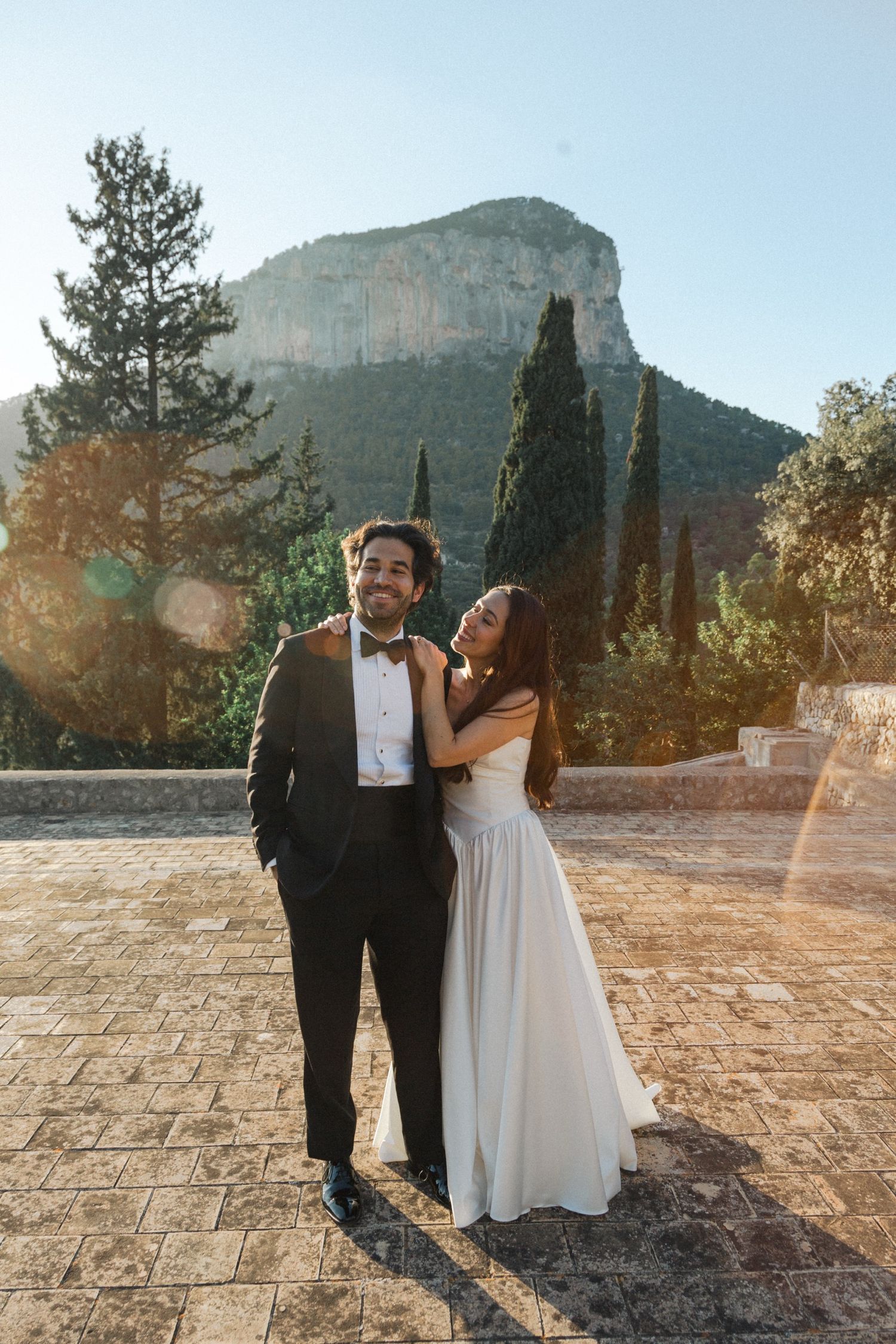 A couple in wedding attire shares a joyful moment together against a scenic mountain backdrop during golden hour.