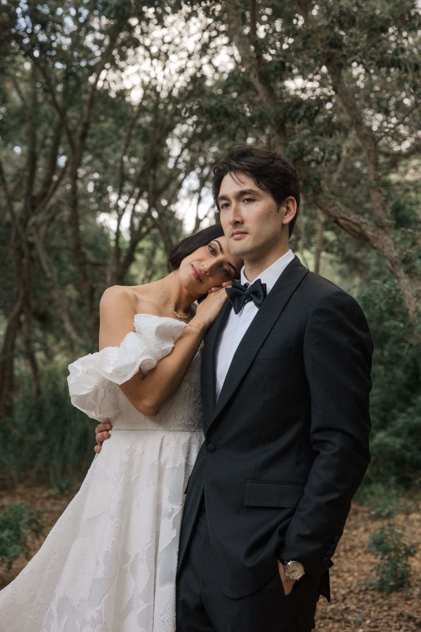 A romantic wedding couple embraces in a wooded setting, the bride in white and groom in classic black tuxedo.