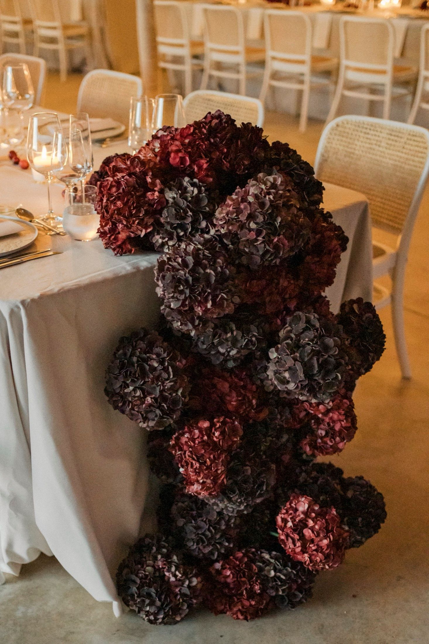Deep burgundy and dark purple hydrangea floral arrangement cascades down a white tablecloth at an elegant dinner setting.