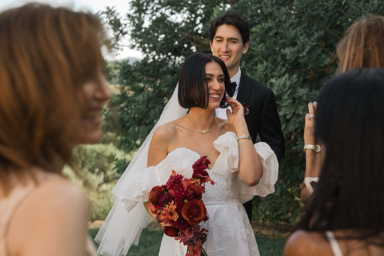 Wedding guests gather outdoors in a garden setting with trees and natural greenery in the background.