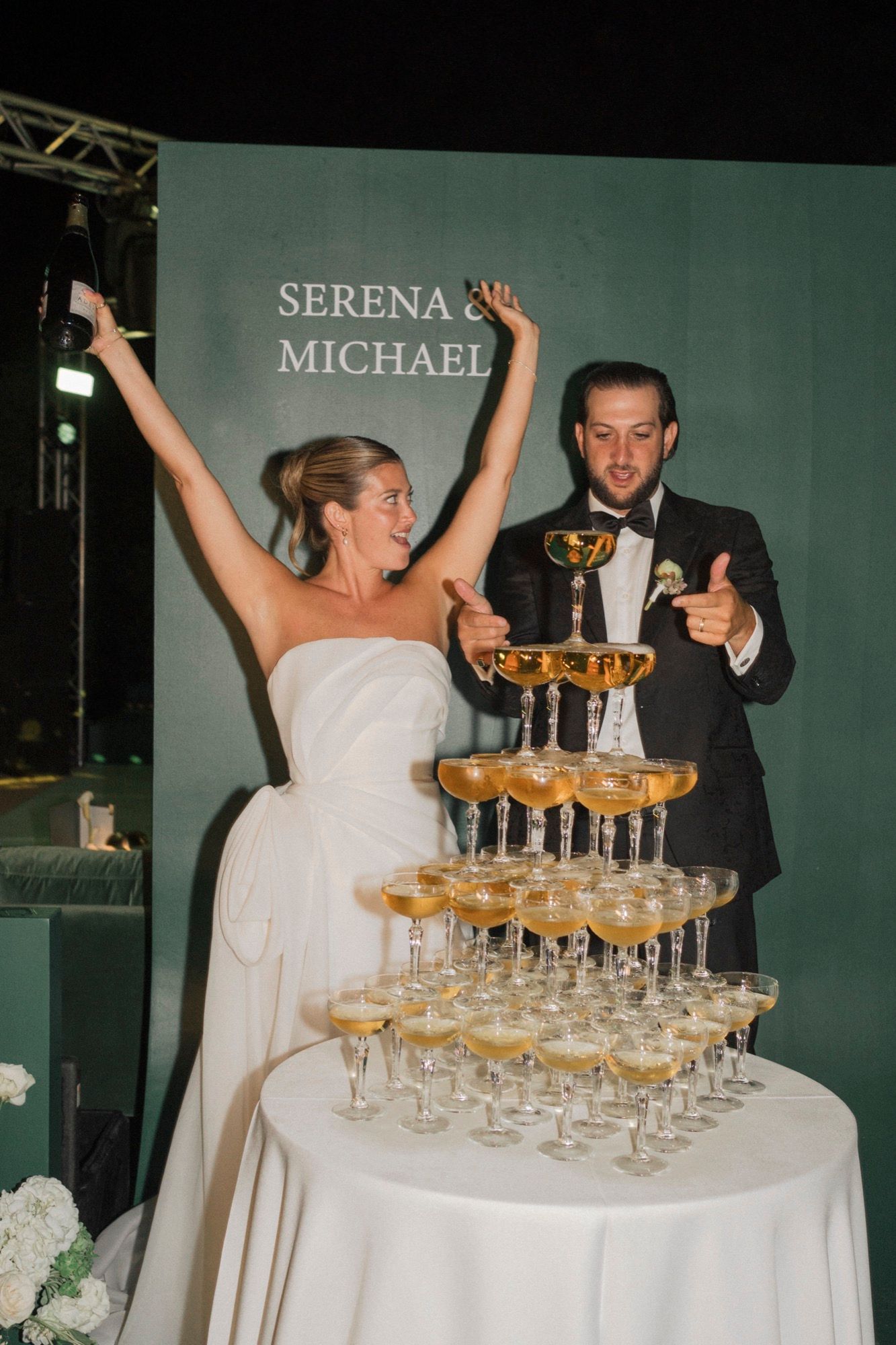 Wedding couple celebrates near a tower of champagne glasses at their reception.