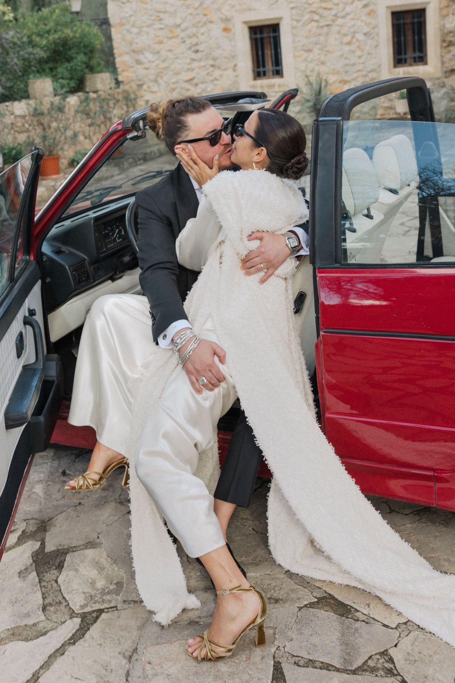 A romantic wedding moment by a vintage red car as a couple embraces in elegant attire.